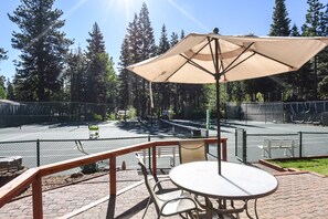 Ein Sitzbereich im Freien mit Tisch und Stühlen, einem großen Sonnenschirm und Blick auf einen von Bäumen umgebenen Tennisplatz.