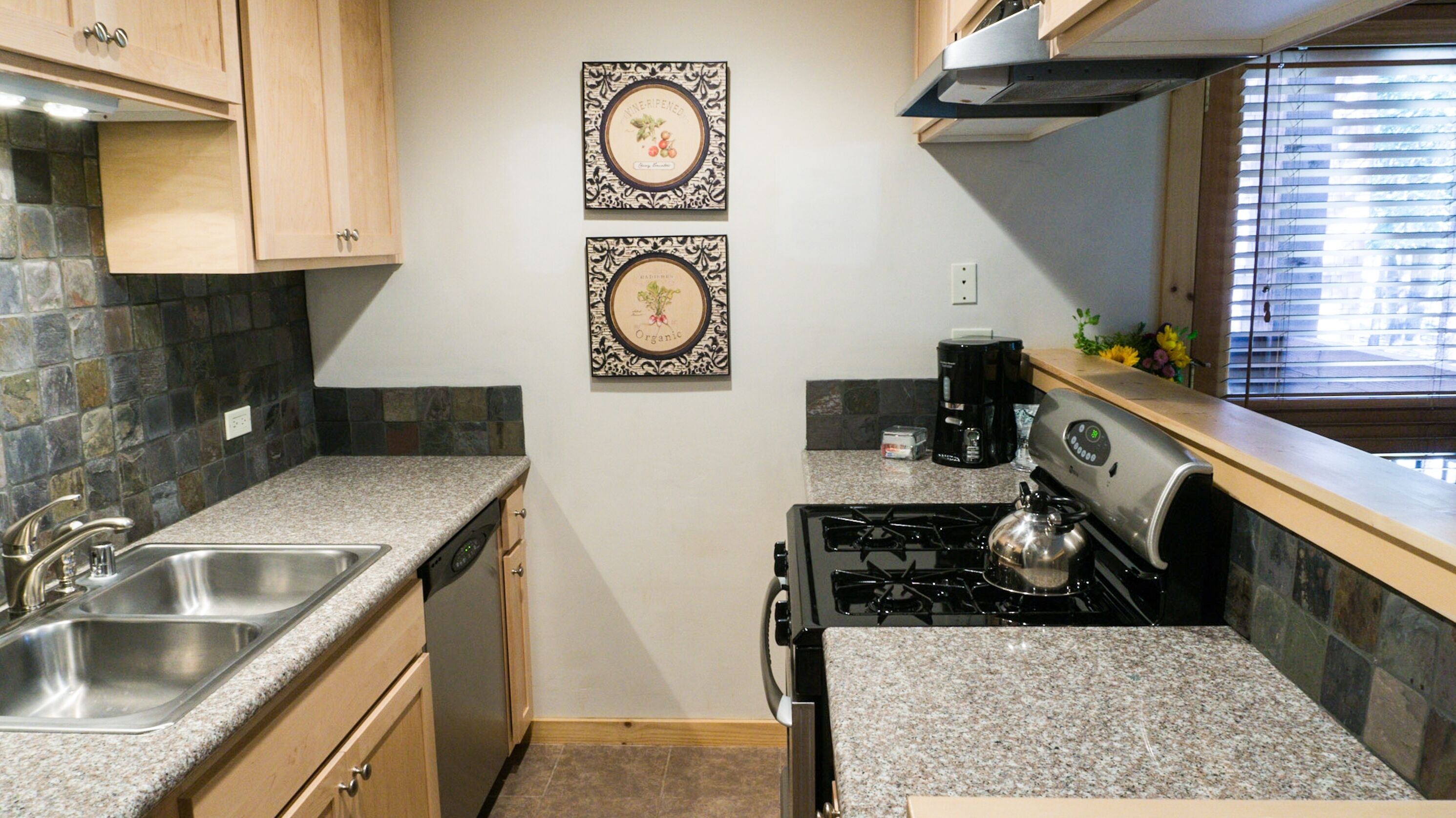 A kitchen with granite countertops, a stainless steel sink, a stove, and two framed pictures on the wall.