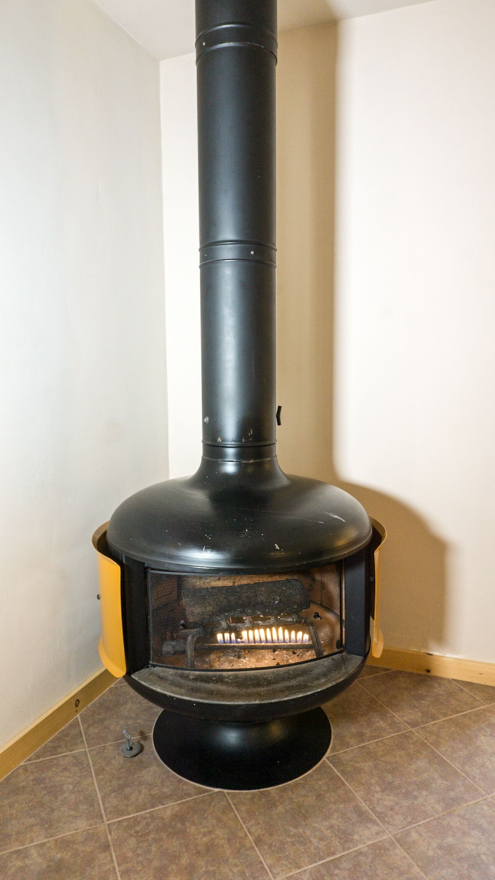 A black wood stove with a glass front, positioned in a corner of a room with tiled flooring and a plain wall.