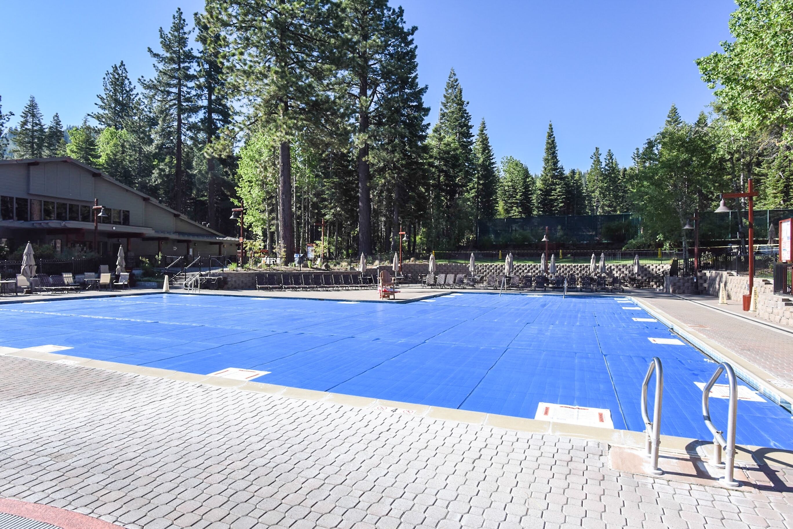 A swimming pool with a blue deck, surrounded by trees and a building with a sloped roof.