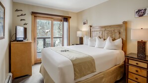 Habitación de hotel con una cama grande, lámparas de noche, un televisor sobre un mueble de madera y un balcón con vistas a árboles.
