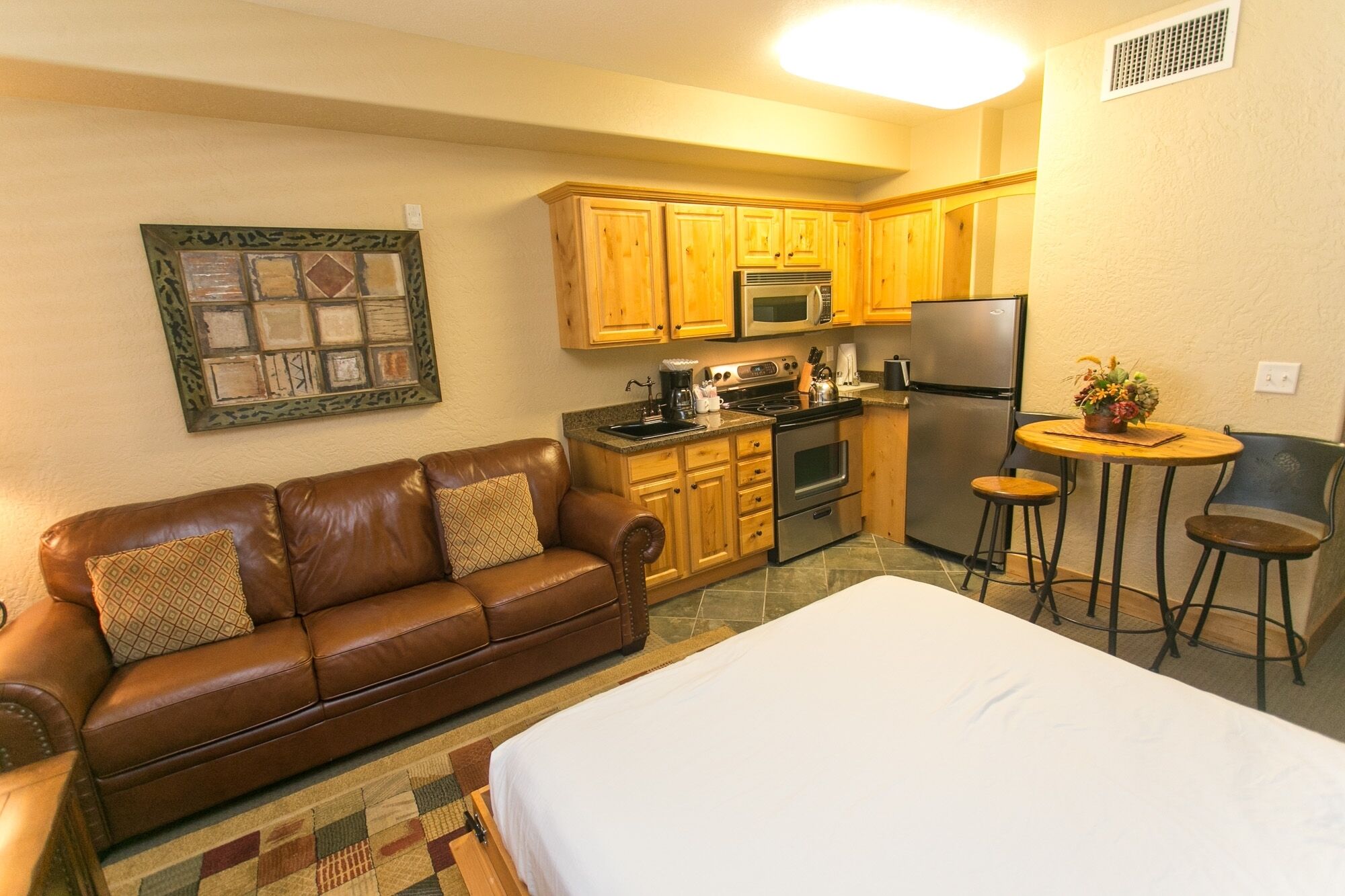 A compact kitchen with wooden cabinets, a refrigerator, and a microwave. There's a brown leather sofa with patterned cushions and a small round table with two stools.