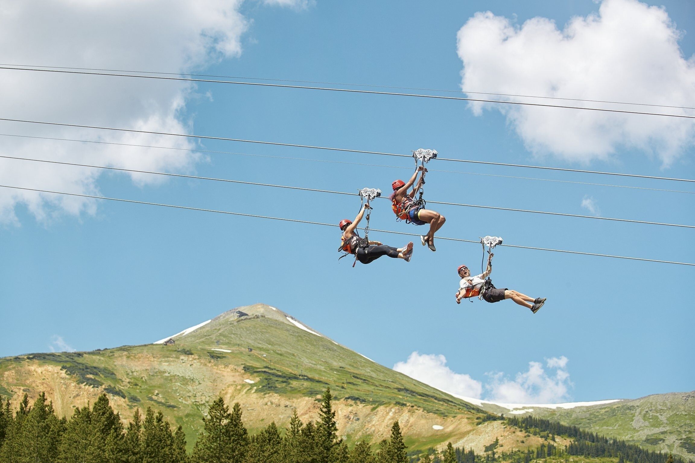 Tres personas suspendidas en el aire, sujetas por cables, con una montaña y cielo despejado al fondo.
