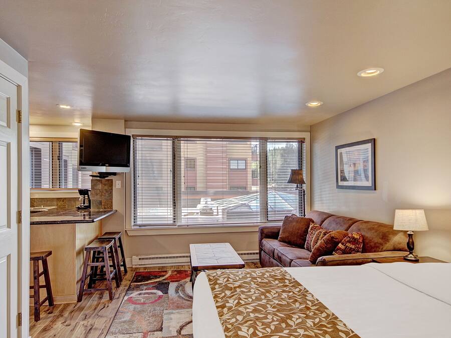 A hotel room with a bed, a sofa, a TV, a small table, and a window with blinds.