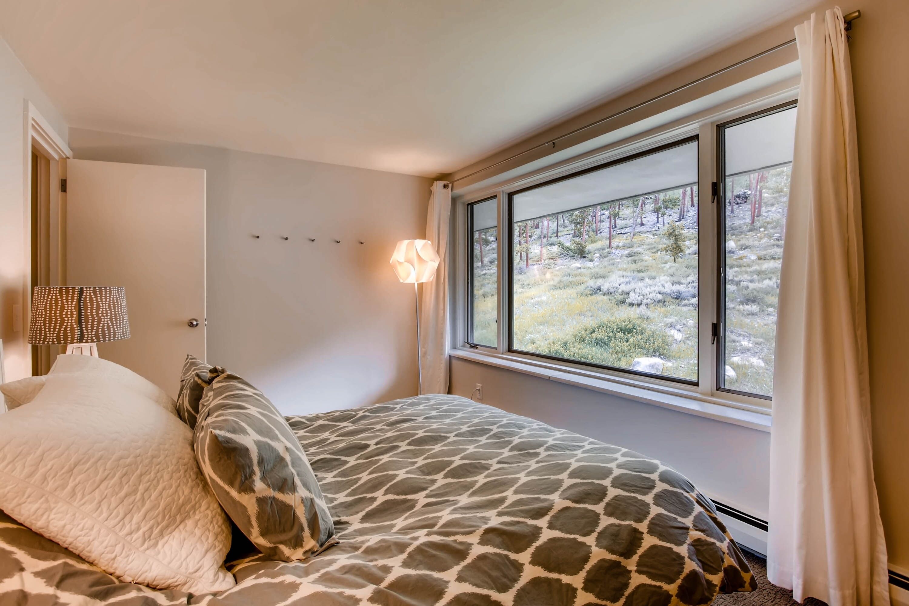 Ein Schlafzimmer mit einem Bett, einer Lampe, einem Fenster mit Blick auf Bäume und einer Tür.