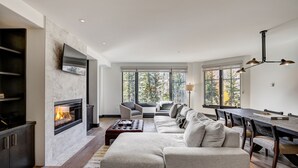 A modern living room with a fireplace, a sectional sofa, a dining table with chairs, and large windows offering a view of trees. - Ski-in/ Ski-out 3br 3.5ba Forest + Mountain Views Sleeps 8 3 Bedroom Condo by RedAwning (Avon)