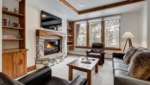 A cozy living room with a stone fireplace, leather sofas, wooden shelves, and a large window offering a view of trees. - Elkhorn 408 (Avon)