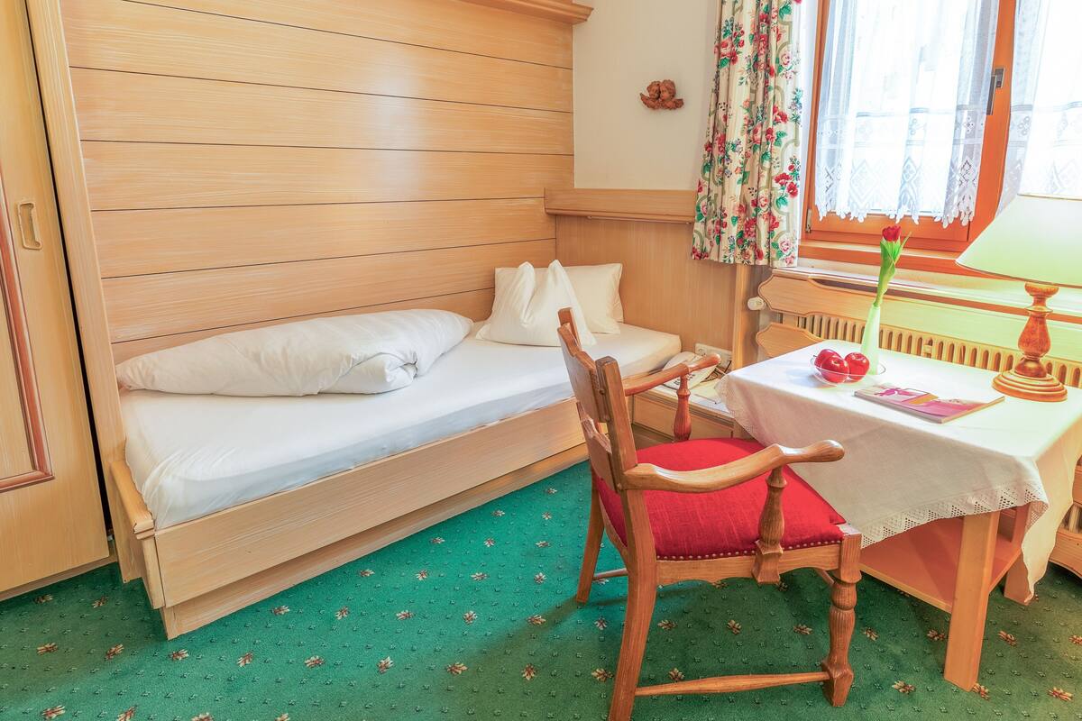 A small, cozy room with a wooden bunk bed, a wooden chair with a red cushion, a small table with a white tablecloth, and a window with floral curtains.