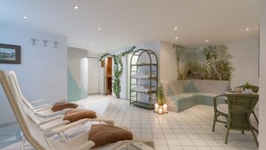 Un área de spa con pisos de baldosas blancas, sillones beige con cojines marrones y una silla de mimbre verde, además de un estante empotrado con toallas.