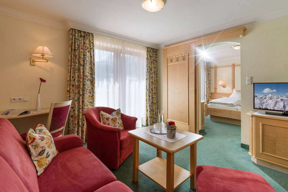 A hotel room with a red sofa, a small table with a vase, a bed with a TV, and a mirror.