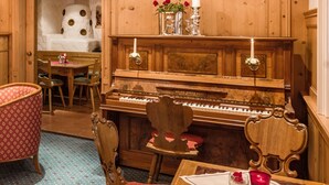 Una habitación acogedora con un piano de cola, sillas de madera, una mesita con un florero y una vela, y una alfombra estampada.