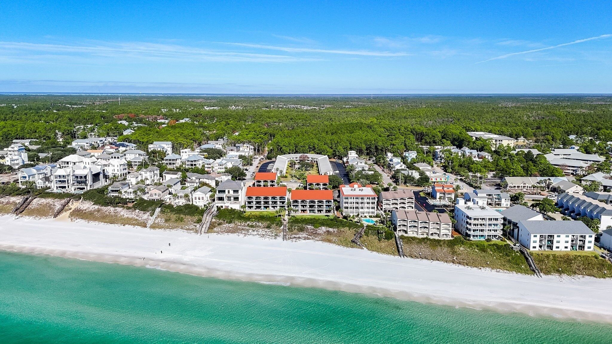 An aerial view of a coastal resort with beachfront properties, a central building with a red roof, and surrounding greenery.