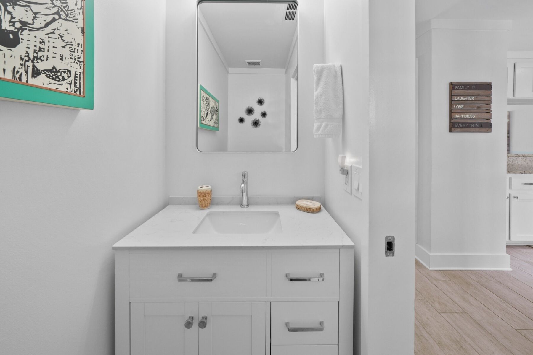 A modern bathroom with a white vanity, a mirror, and a small sink. There is a framed picture on the wall and a sign with words like "family" and "love".