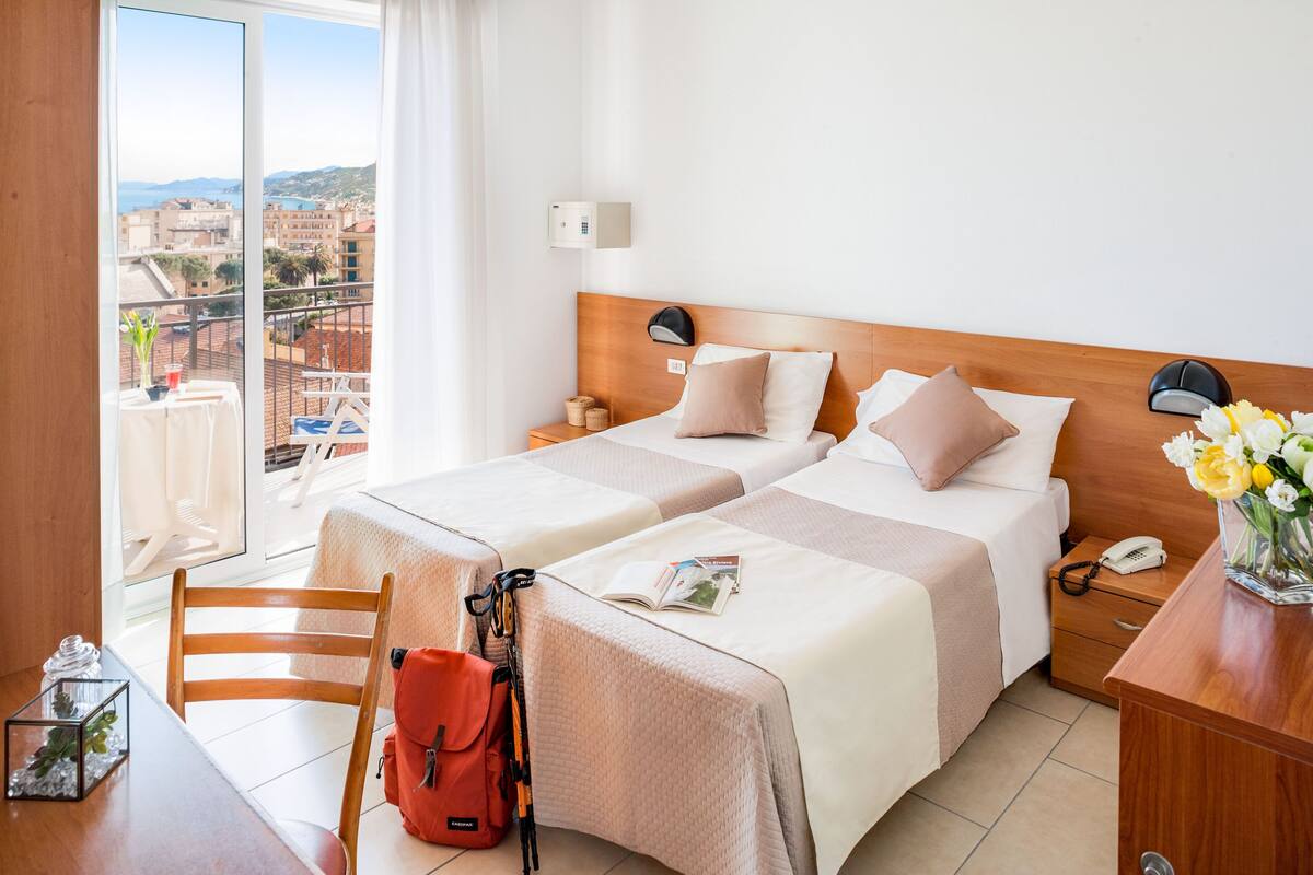 Une chambre d’hôtel avec deux lits, une tête de lit en bois, une table de chevet avec un téléphone, une table avec une chaise, et une vue sur la ville.