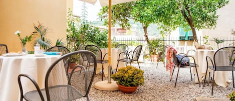 Un área de comedor al aire libre con mesas y sillas, rodeada de plantas en macetas y árboles.