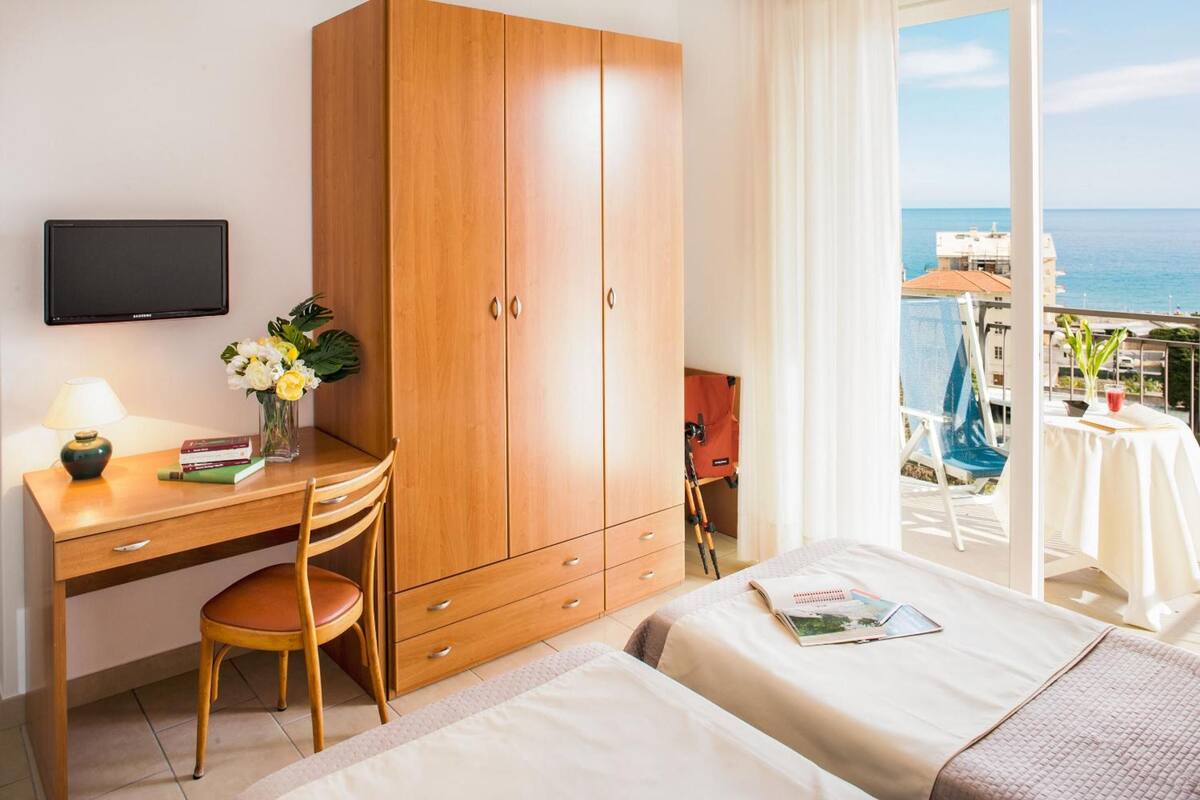 Une chambre d’hôtel avec un bureau en bois, une chaise, un lit, une télévision et un balcon avec vue sur la mer.