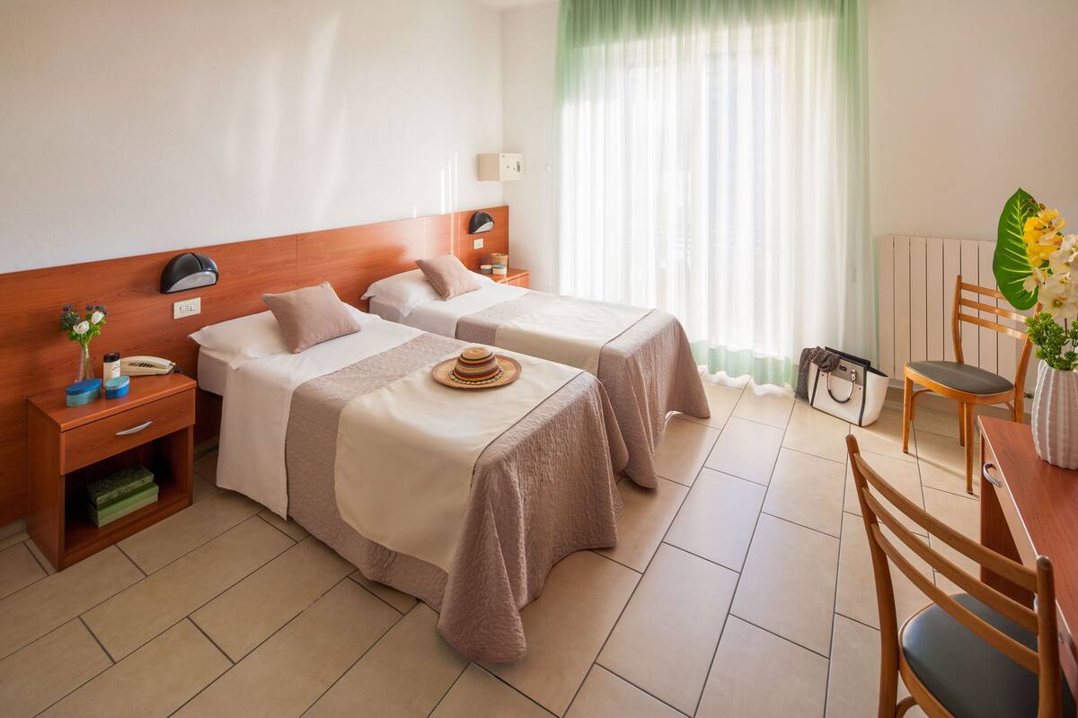 Une chambre d’hôtel avec deux lits, une tête de lit en bois, des tables de chevet avec des lampes, une table à manger avec des chaises et une grande fenêtre avec des rideaux légers.
