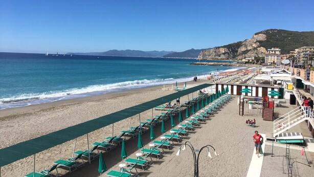 A beach with rows of sun loungers and umbrellas, a promenade, and a view of the sea and distant mountains. - Hotel Europa (Finale Ligure)