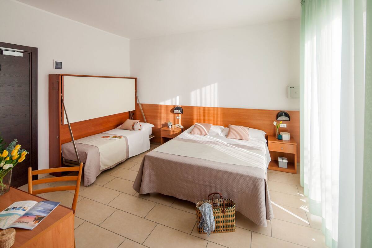 Une chambre d’hôtel avec deux lits, un bureau avec une chaise, une petite table ornée d’un vase de fleurs et une table de chevet avec des lampes.