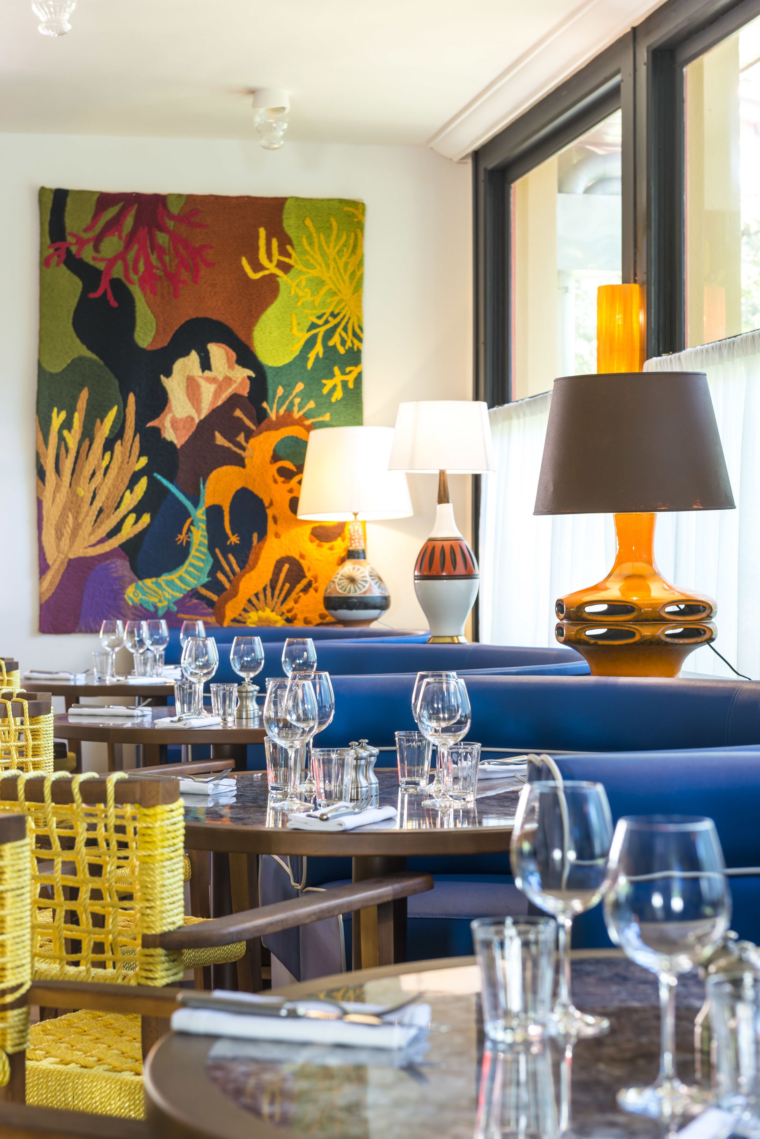 Een restaurant met een blauwe bank, meerdere tafels met wijnglazen en een kleurrijk schilderij aan de muur.