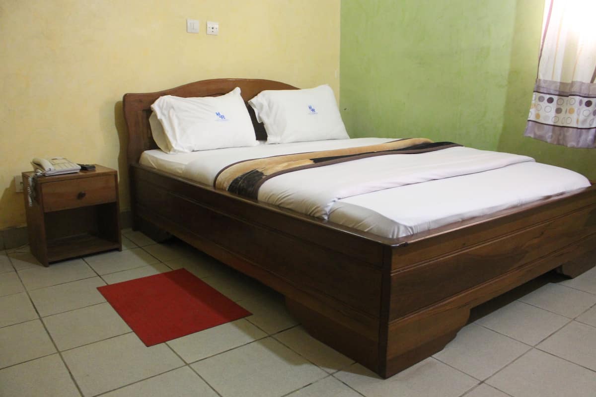 Una cama individual con estructura de madera, ropa de cama blanca y una alfombra roja sobre suelo de baldosas.