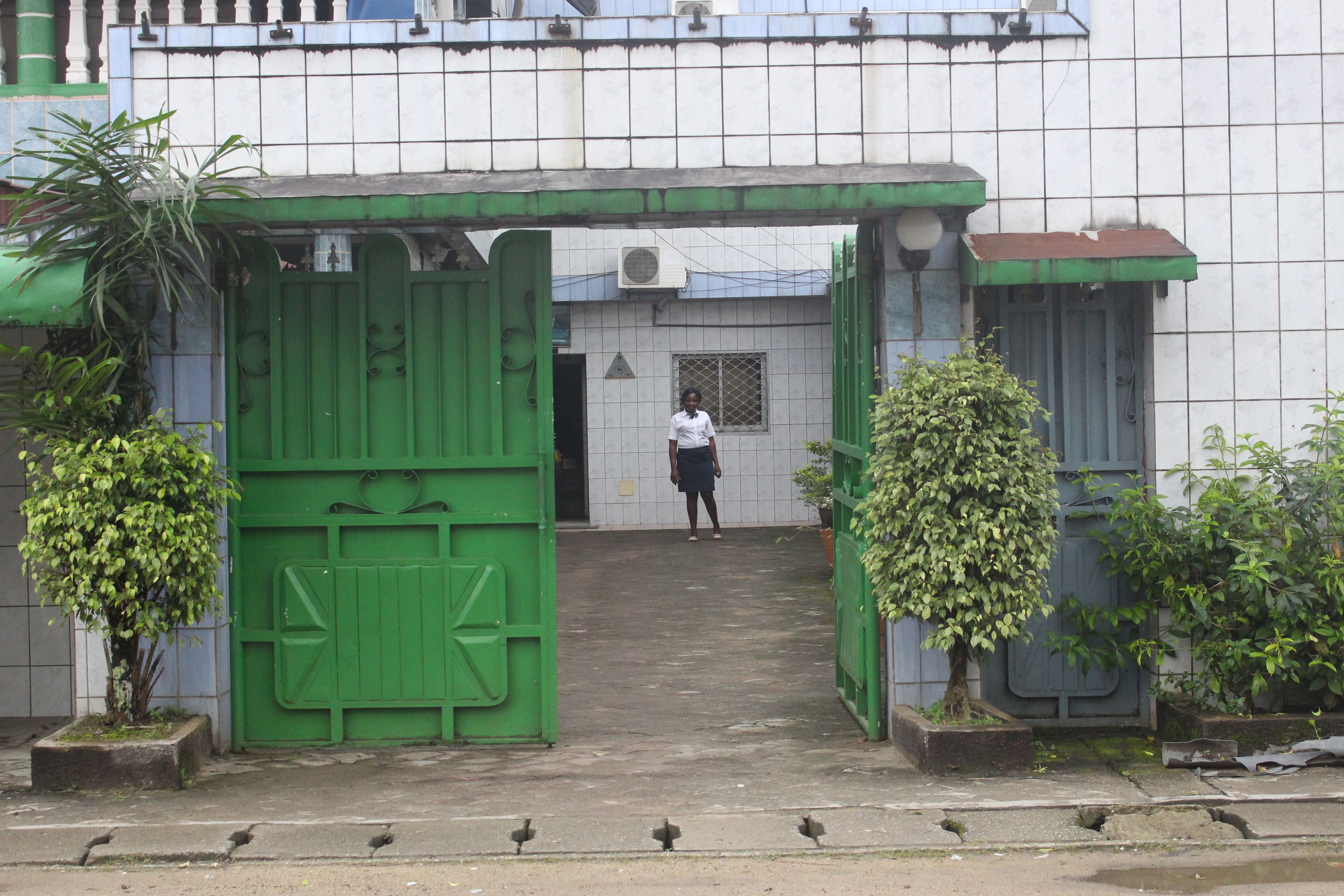 A gated entrance with a person standing in the distance, surrounded by greenery and a tiled wall.