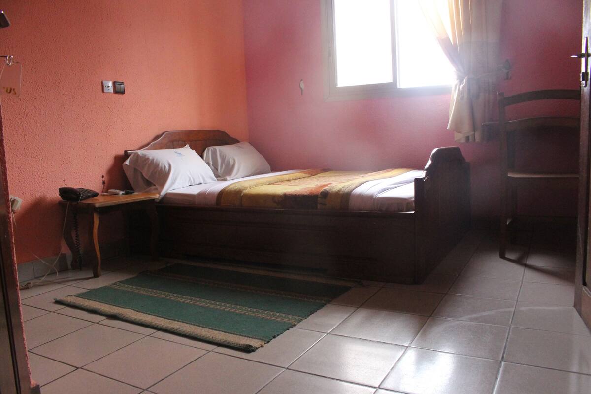 Una cama individual con estructura de madera, una mesita de noche con lámpara, una alfombra verde y una ventana con cortinas.