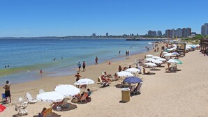 Une plage avec des transats et des parasols, une jetée en arrière-plan et une vue sur les gratte-ciel de la ville de l’autre côté de l’eau.
