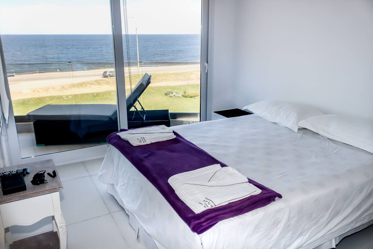 A hotel room with a large window offering a view of the beach and ocean. The room has a bed with white linens, a purple throw blanket, and a neatly made bed.