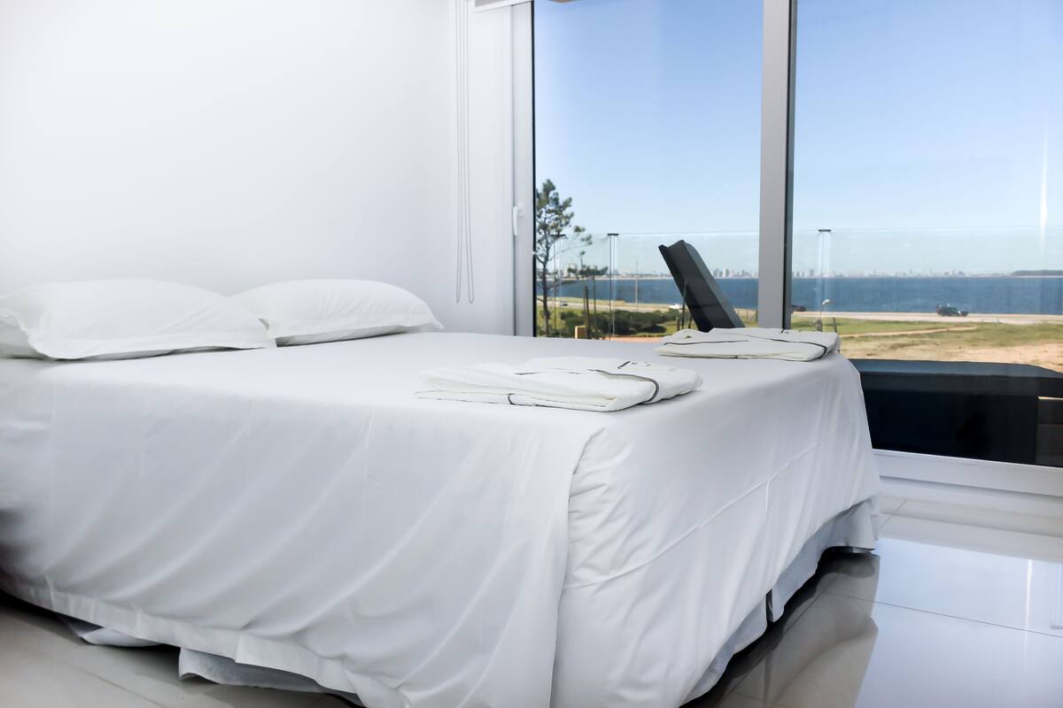 A modern bedroom with a large bed, white linens, and a view of the ocean through floor-to-ceiling windows.