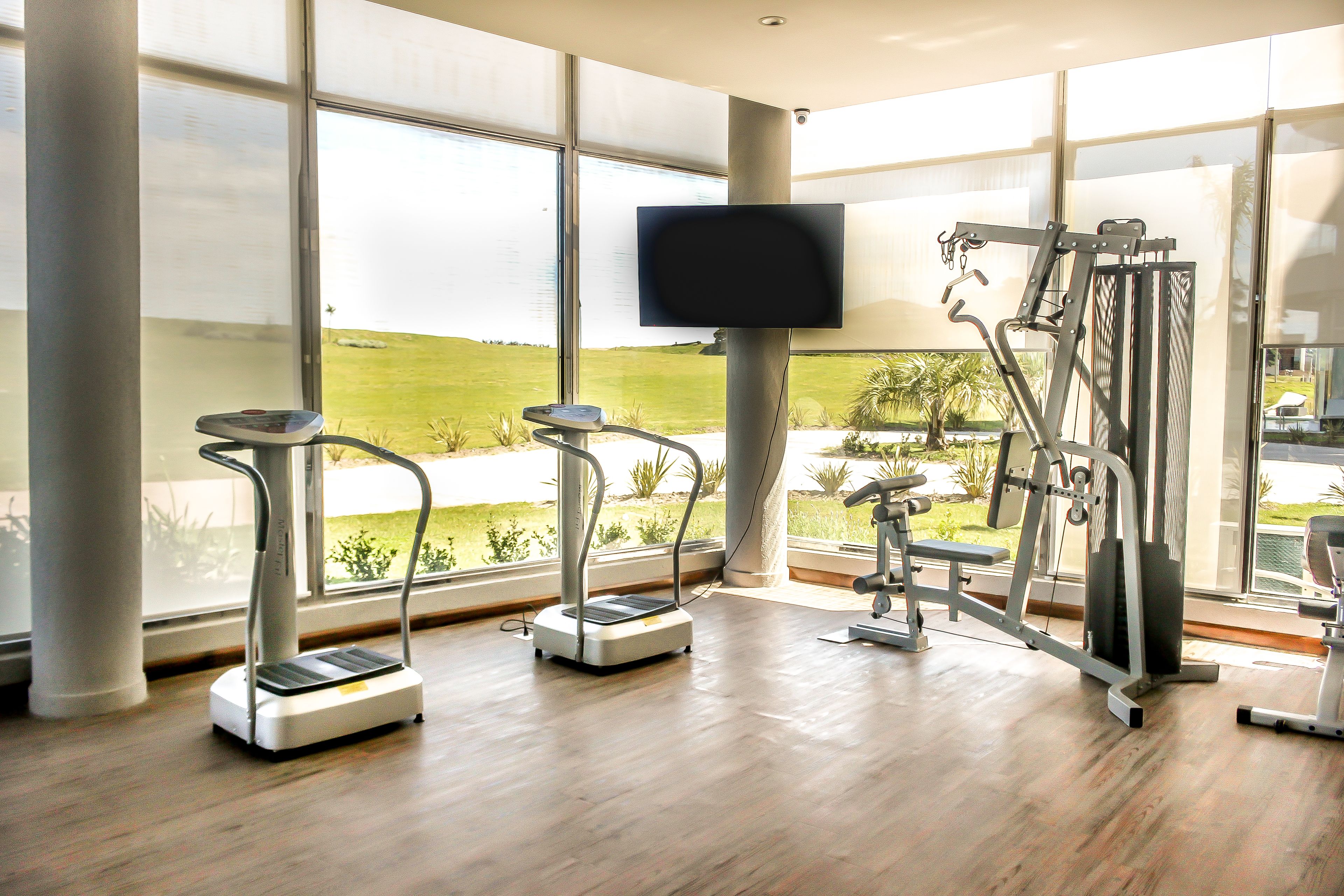 A gym with treadmills, a stationary bike, and a weight machine, large windows, and a TV on a pillar.
