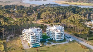 Un immeuble résidentiel moderne à plusieurs étages, doté d’une cour intérieure, entouré d’une forêt dense et avec un plan d’eau en arrière-plan.