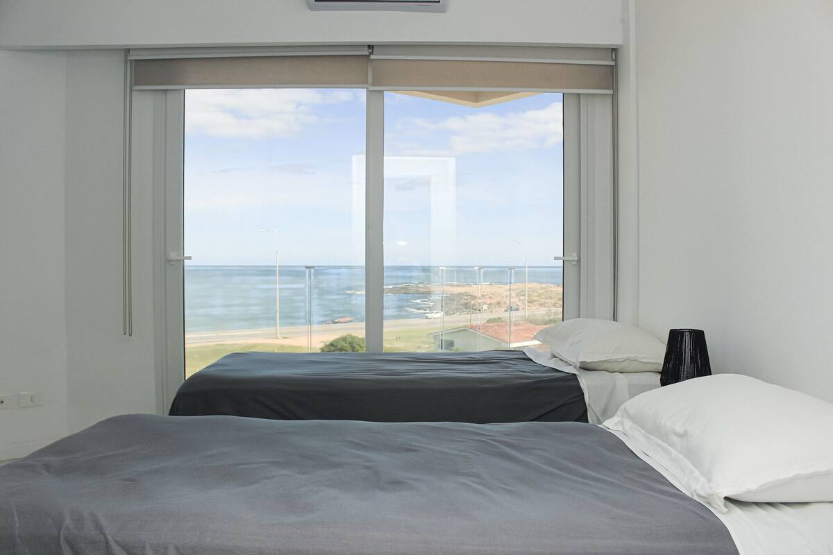 A bedroom with two beds, a large window offering a view of the sea and a distant landscape.