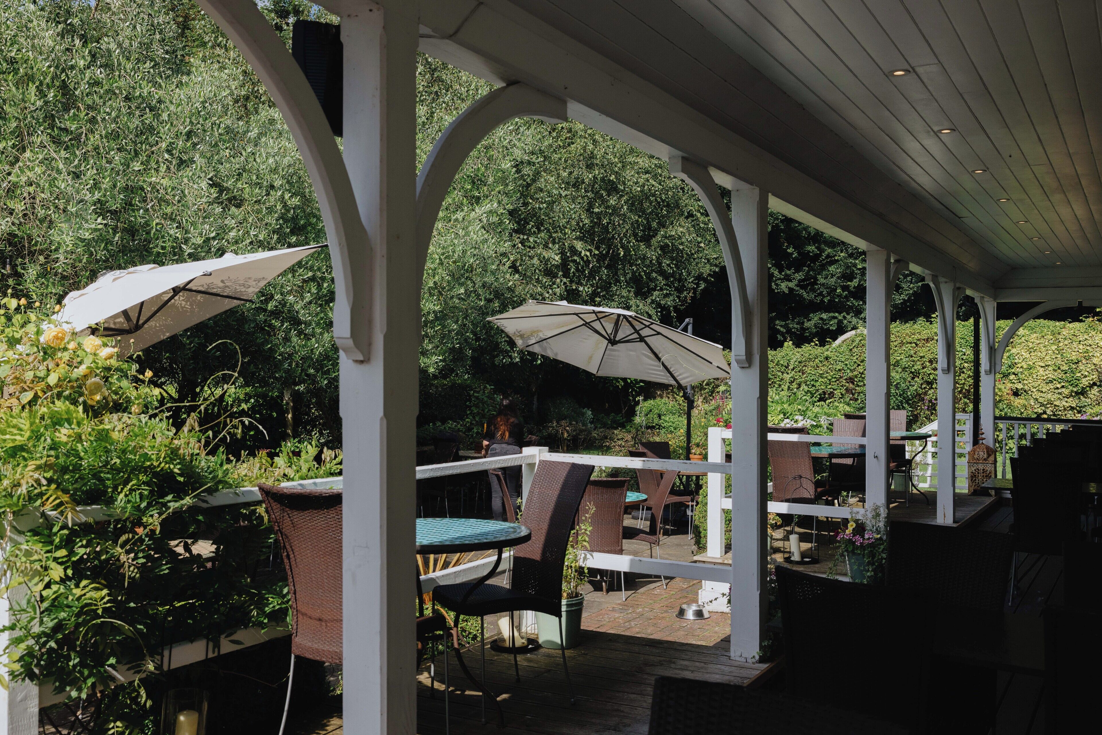 An outdoor patio area with white columns, round tables, and chairs, surrounded by greenery.