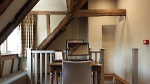 Ein Zimmer mit einem Holztisch, einem Stuhl und einem Spiegel. Ein Fenster mit Vorhängen bietet Blick ins Freie. Die Decke ist schräg und mit Holzbalken versehen.