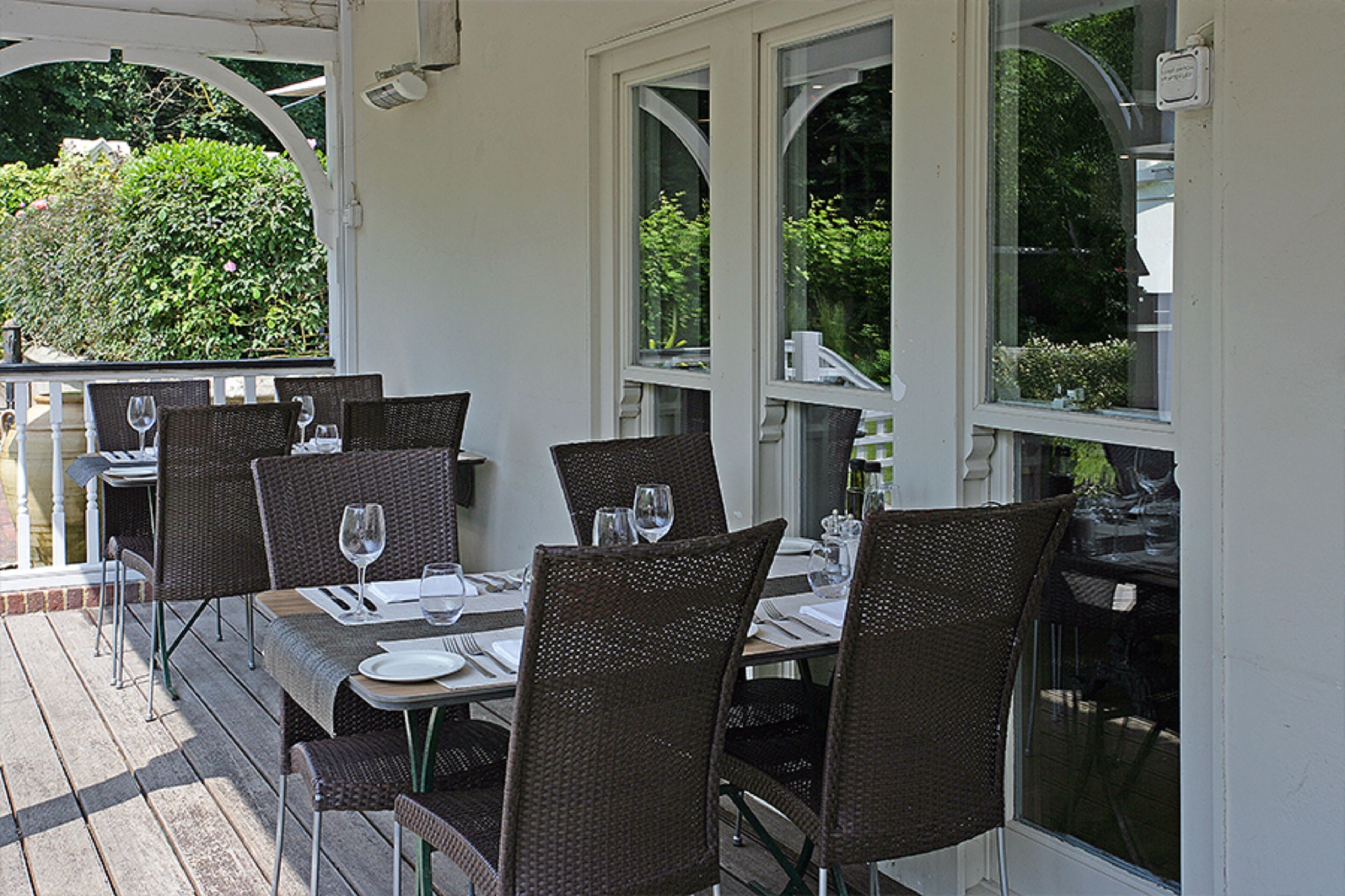An outdoor dining area with wicker tables and chairs, white walls, and large windows overlooking greenery.