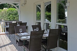 An outdoor dining area with wicker tables and chairs, white walls, and large windows overlooking greenery.