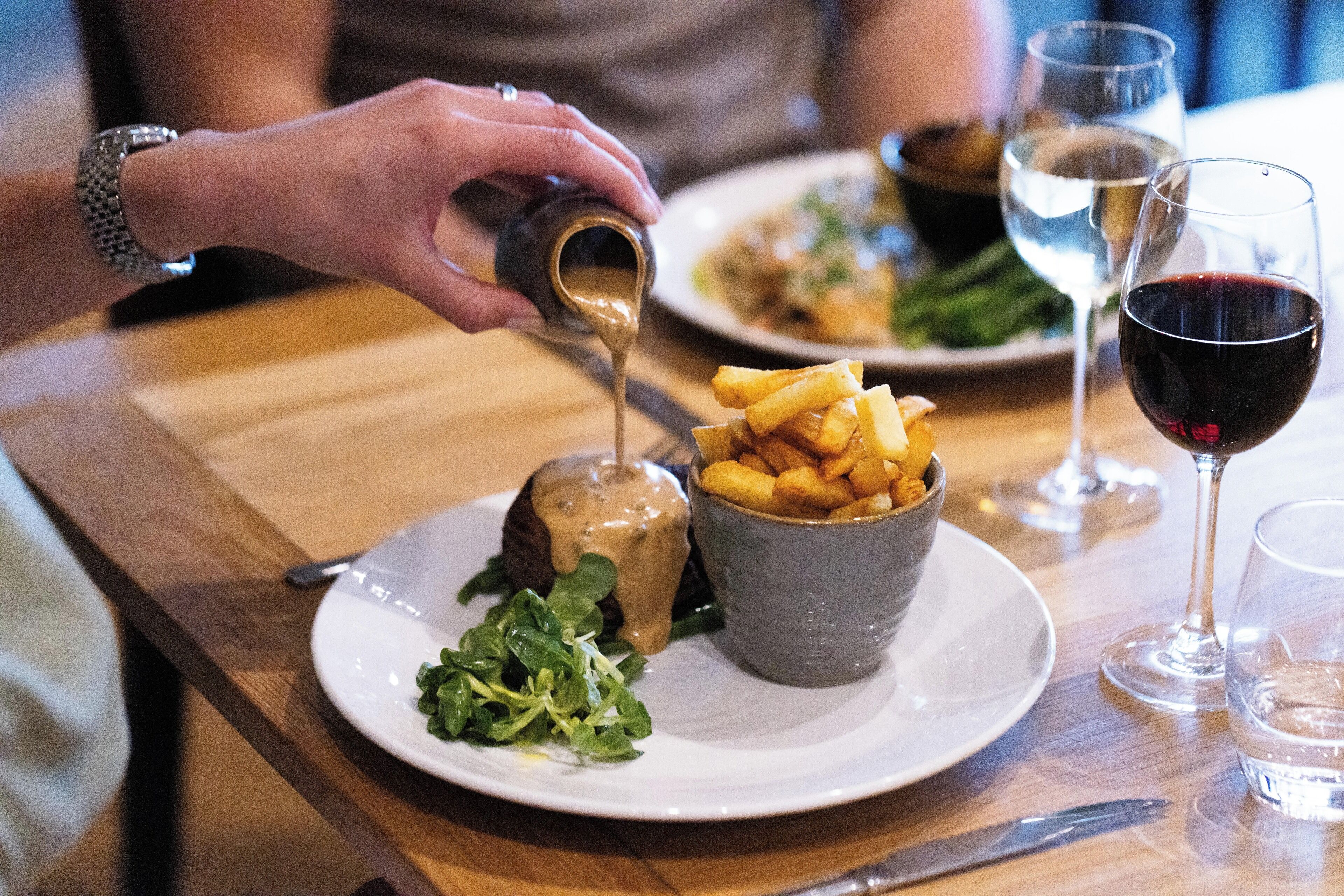 Una mesa de comedor con un plato de comida, un bol de papas fritas y una copa de vino tinto.