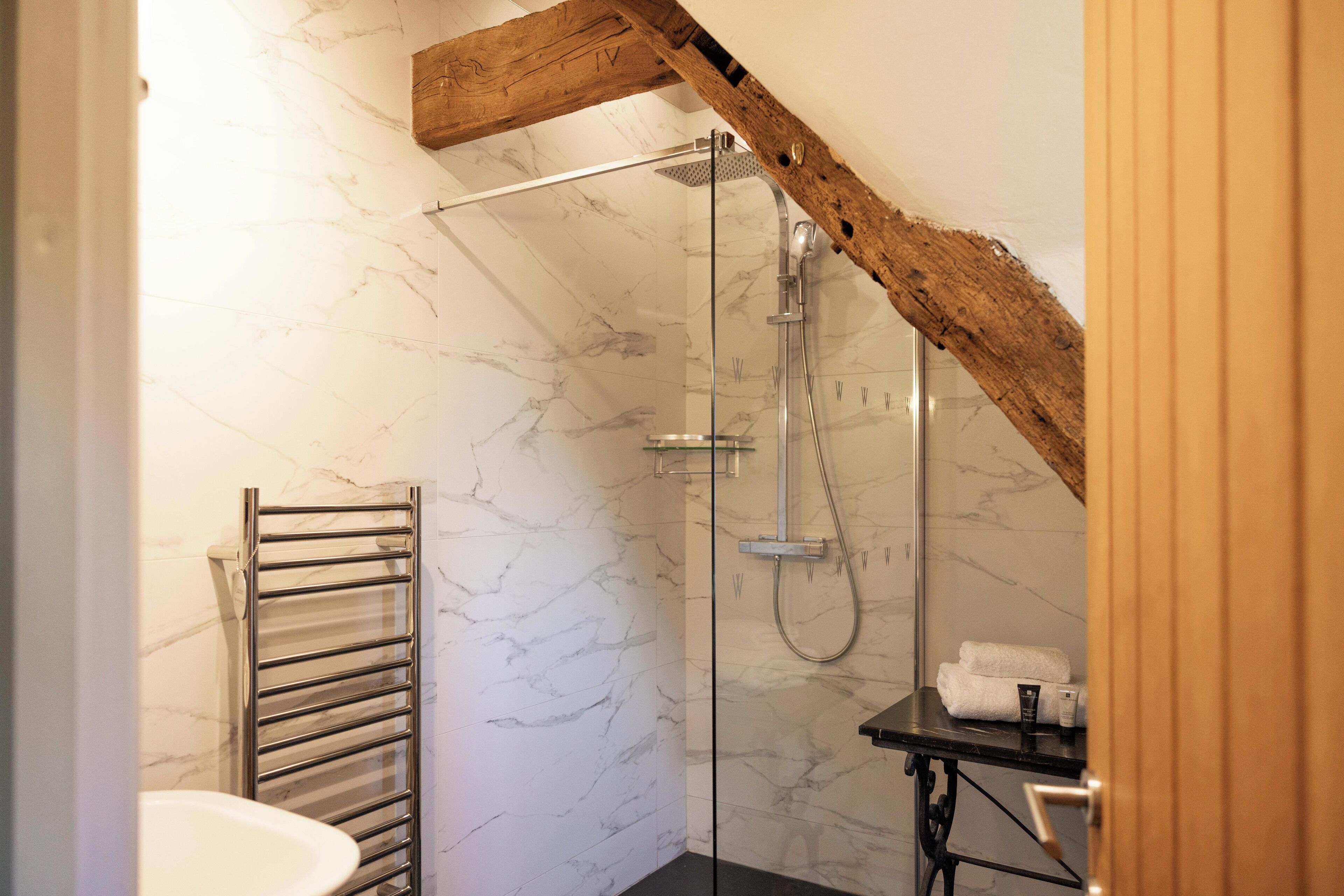 A modern bathroom with a glass-enclosed shower, marble walls, and a wooden ceiling beam.
