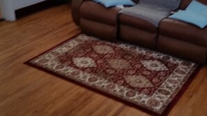 A living room with a brown leather sofa, a patterned rug, and wooden flooring. - Just You in Neepawa Vacation Home (Neepawa)