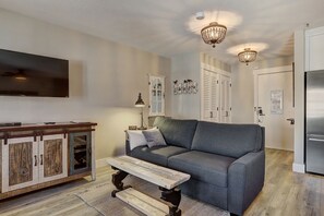 A living room with a grey sofa, wooden coffee table, and a cabinet with glass doors. There are two chandeliers, a flat-screen TV, and a door with a lock.