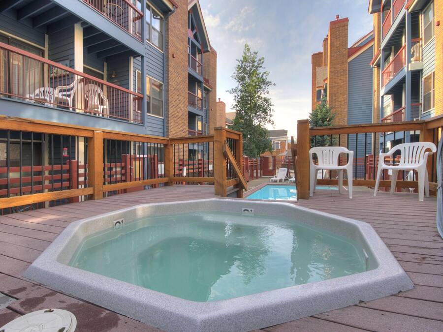 A hot tub on a wooden deck with a pool in the background, surrounded by buildings with balconies and outdoor seating.