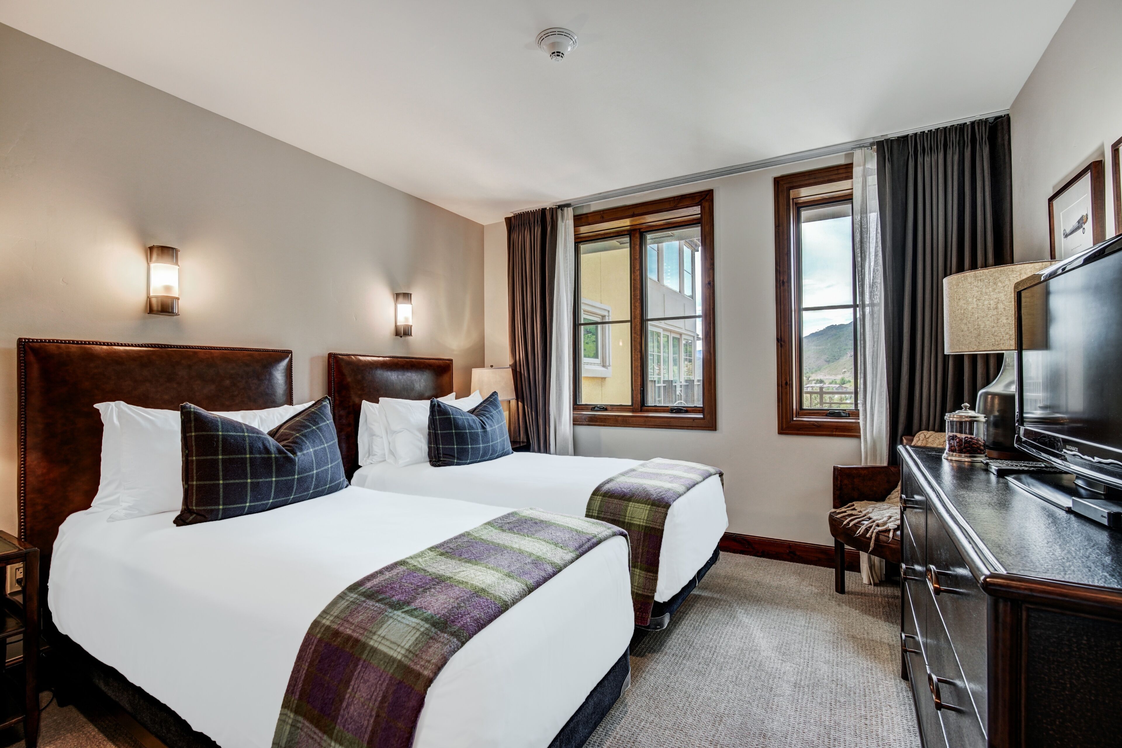 A hotel room with two beds, a desk with a TV, and a window with curtains.