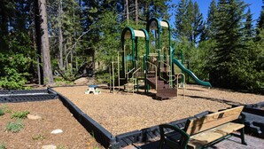 Ein Spielplatz mit Rutsche und Klettergerüsten, umgeben von Bäumen und einer gepflasterten Fläche mit einer Bank.