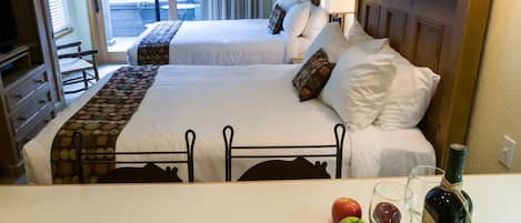A hotel room with two beds, a desk with a bottle of wine, two apples, and two wine glasses.