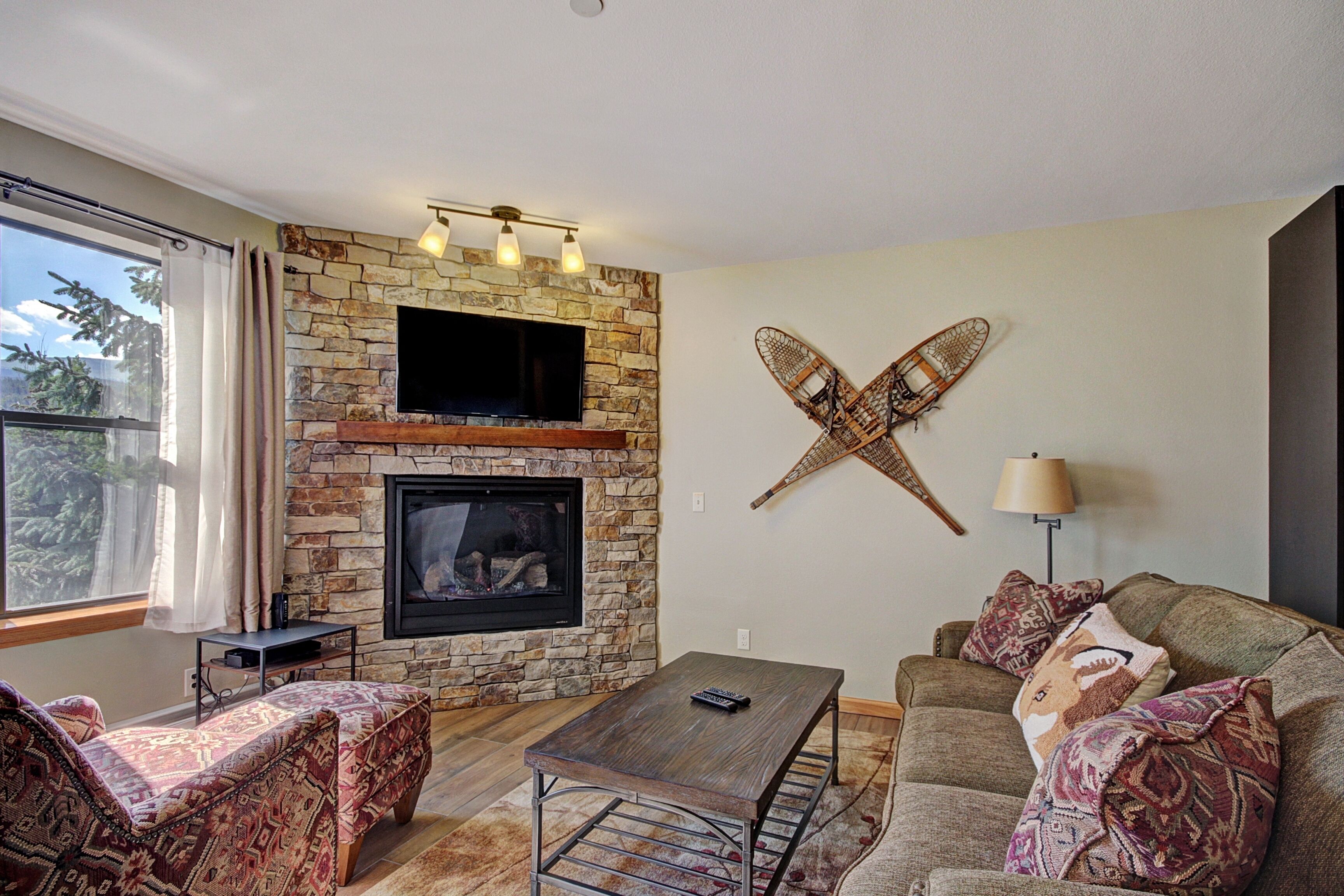 A living room with a stone fireplace, a flat-screen TV, a wooden coffee table, patterned sofas, and a large window with curtains.