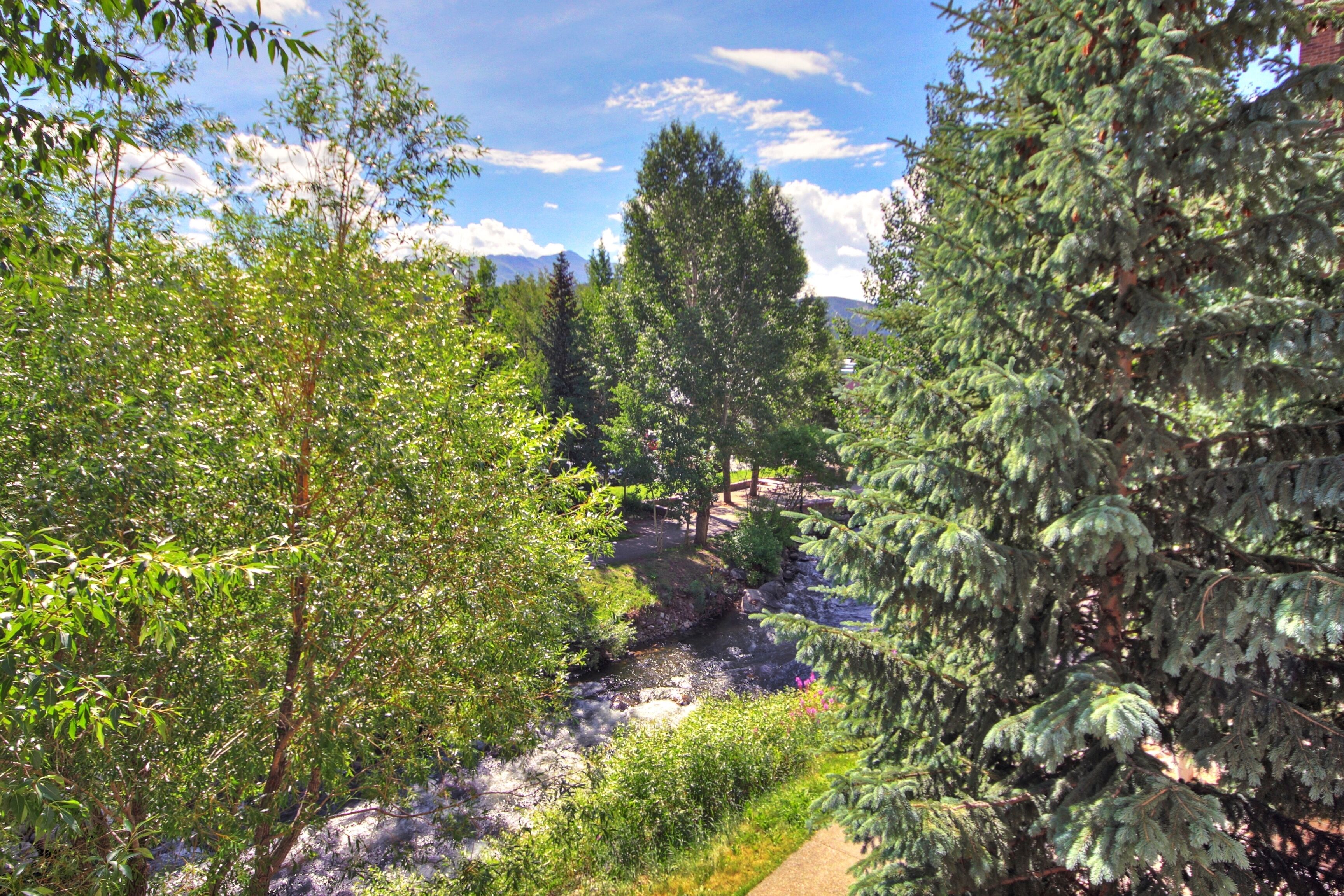 A natural landscape with a stream, dense trees, and a clear sky.