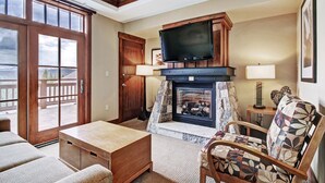 A cozy living room with a fireplace, a television mounted above it, and a large window offering a view of the outdoors. - Luxury Studio  Steps From Winter Activities, Sleeps 4! Studio Bedroom Condo by RedAwning (Breckenridge)