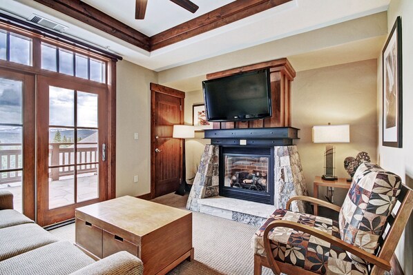 A cozy living room with a fireplace, a television mounted above it, and a large window offering a view of the outdoors.