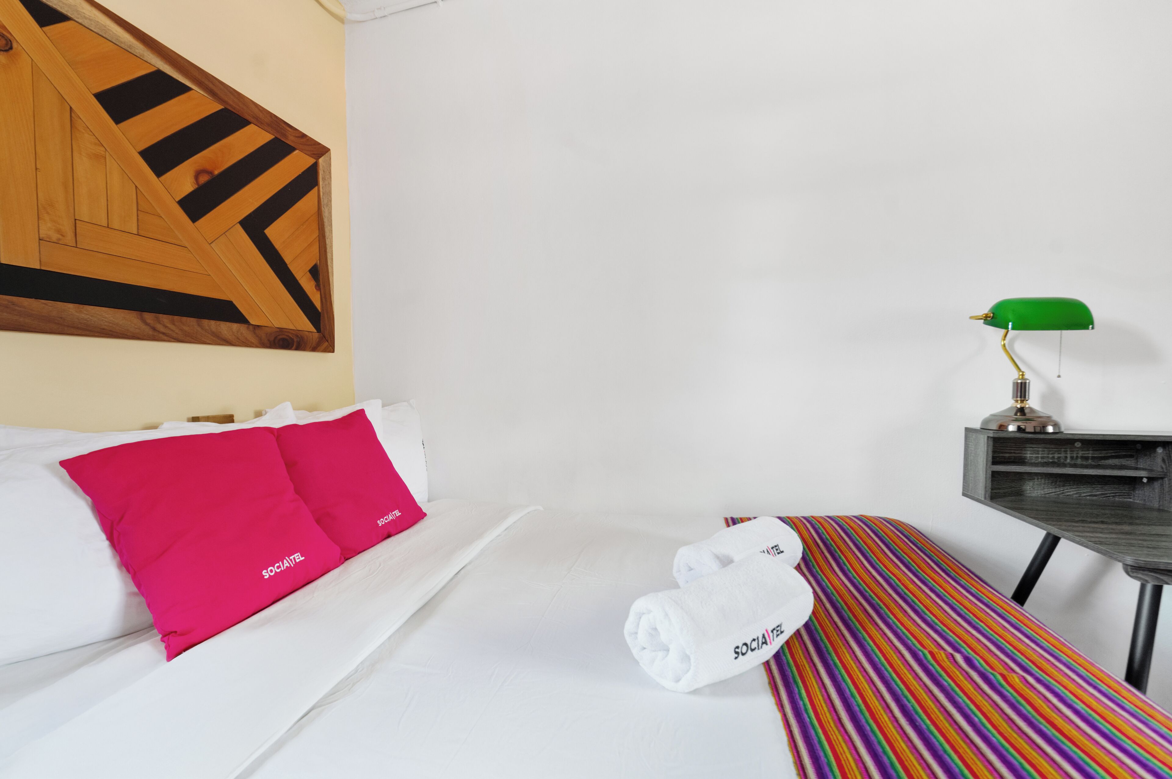 A hotel room with a bed, a bench, a green desk lamp, and a patterned wall.